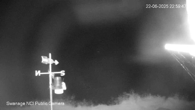 A black-and-white image from a webcam shows a weather vane with an arrow pointing east and a cylindrical device attached to it. The background is dark, with some blurry vegetation at the bottom. The timestamp in the top right corner reads "22-06-2025 22:59:47," and there is white text at the bottom left that says "Swanage NCI Public Camera."