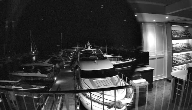 A black-and-white image taken from a webcam shows a marina at night with several boats docked. In the foreground, there is a large boat with a sleek design, placed indoors, possibly a showroom or an enclosed space. To the right, there's a wall with shelves displaying what appears to be model boats or nautical items. A round table is partially visible in the lower right corner, and the ceiling has recessed lighting.