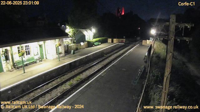 A quiet, empty railway station platform at night, illuminated by a few bright lights. The platform has a bench, a small building with flower boxes, and some signboards. Railway tracks run alongside the platform, and there is a utility pole with wires on the right side of the image. The sky is dark, and the overall scene appears calm and still.