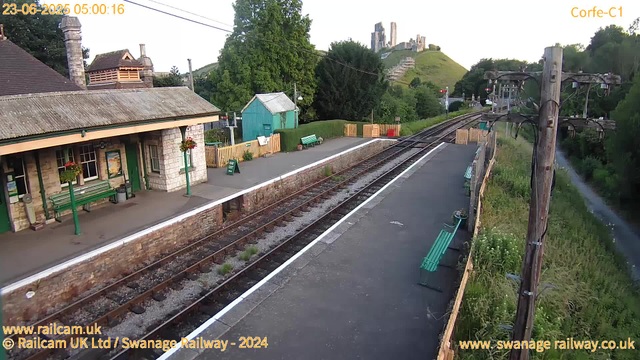 A small railway station platform with a stone building on the left, decorated with hanging flower baskets. There are green benches and a small waiting area, with a "Way Out" sign on a green post. To the right, there are railway tracks with a wooden fence along the edge of the platform. In the background, a hillside features the ruins of an ancient castle or fortress. Tall trees and lush green vegetation surround the scene, and the early morning sky is clear. The timestamp in the top left indicates it was taken at 5:00 AM on June 23, 2025.