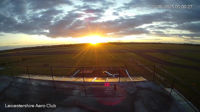 The image shows the sunrise at Leicestershire Aero Club with the sun just above the horizon, casting bright rays across the sky. There are scattered clouds with some darker, heavier clouds on top and a wide open grassy airfield area with a small section of a runway visible on the right. The foreground includes a partially visible rooftop and safety railings. The timestamp indicates the photo was taken on June 23, 2025, at 5:00 a.m.
