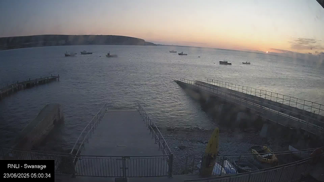 A view of a harbor at sunrise, with several small boats floating on calm water. In the foreground, there is a promenade with railings and a small yellow vehicle parked near the edge. A breakwater extends into the water on the right side, and a rocky shoreline is visible at the bottom. The sky is partly cloudy with the sun just beginning to rise on the horizon.