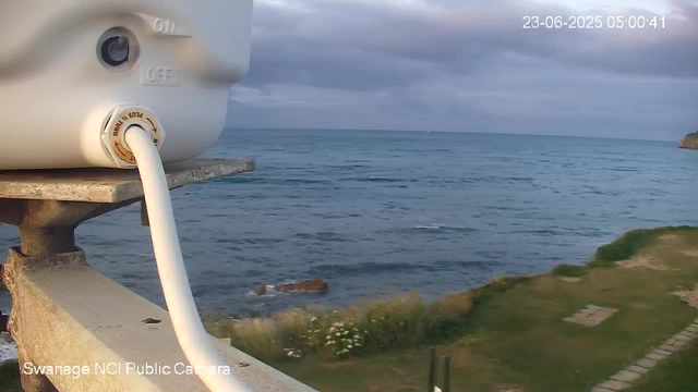 A view of the ocean with a cloudy sky overhead, seen from a webcam mounted on a stone railing. Part of a white telescope or observation device is visible on the left side of the image, connected by a white cable. In the distance, there is a faint silhouette of land or a cliff on the right edge. The timestamp on the top right indicates the date and time as 23-06-2025 05:00:41. The bottom left corner has a label reading "Swanage NCI Public Camera."