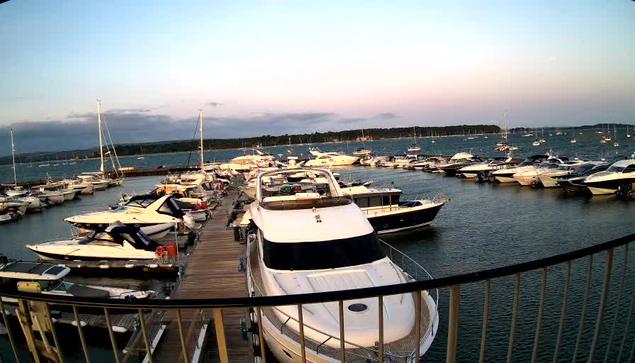 A marina with numerous boats and yachts docked along wooden piers. The water is calm, and the sky above is partly cloudy with a hint of pinkish hue near the horizon, indicating either sunrise or sunset. A safety rail runs along the foreground of the image.