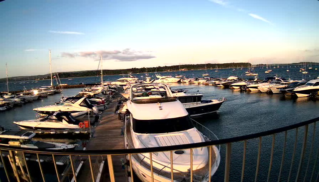 A marina filled with various boats and yachts docked along wooden piers, with a calm body of water and a distant forested shoreline under a partly cloudy sky during sunset.