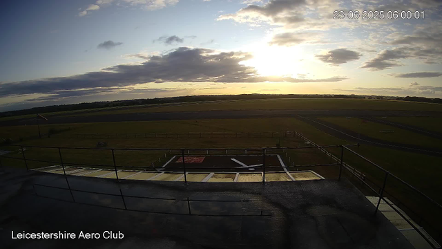 An outdoor scene at sunrise or sunset showing a wide view of a grassy airfield with runways stretching into the distance. The sky is partly cloudy with the sun low on the horizon, casting a warm light. In the foreground, there is a viewing platform with metal railings and a helipad with a yellow circle and markings. The location is identified as Leicestershire Aero Club.