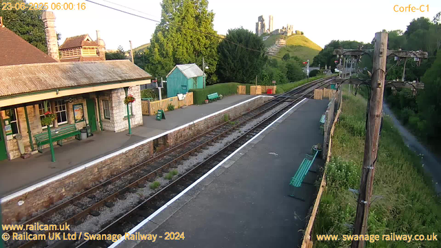 A small train station platform is shown with a building on the left that has a stone exterior, green benches, and hanging flower baskets. The platform has a white safety line and a black top surface. There are a few green benches, a small green shed, and a yellow fence at the far end. To the right, there are overhead electrical wires and a pole. In the background, there are lush green trees, a hill with a castle ruin on top, and a clear sky. The timestamp indicates the date and time as 23-06-2025 at 06:00:16.