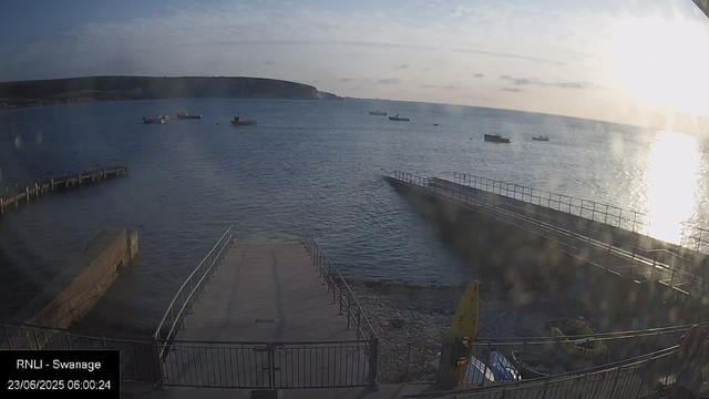 A wide shot of a harbor taken in the early morning. Several boats are anchored in the water, with a coastline and cliffs visible in the distance. The sun is rising on the right side, casting bright light over the scene and creating reflections on the water. In the foreground, there are wooden and metal structures, including ramps and barriers, leading into the water. The timestamp indicates the image was captured on June 23, 2025, at 6:00 AM.