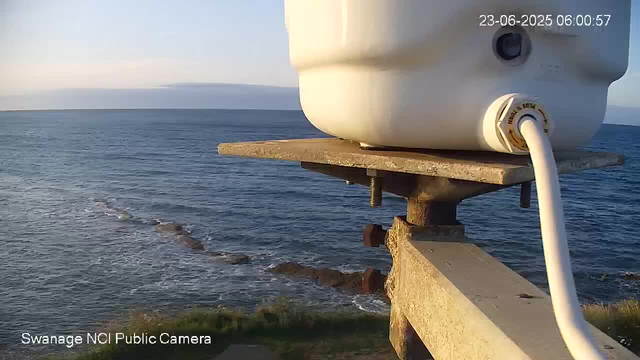 A webcam view overlooking the ocean with gentle waves and a distant horizon. In the foreground, there is a white outdoor camera mounted on a concrete structure, with a cable connected to it. The timestamp shows the date as June 23, 2025, and the time as 06:00:57. The bottom left corner has the label "Swanage NCI Public Camera."
