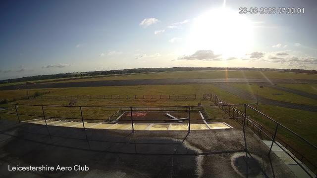 A wide view of an airfield with a grassy area in the foreground, surrounded by a wooden fence. The runway stretches across the middle distance under a partly cloudy sky, with the sun low in the sky, creating a bright glare. A small flag and a few structures are visible near the fence, and there is a timestamp in the upper right corner reading "23-06-2025 07:00:01."