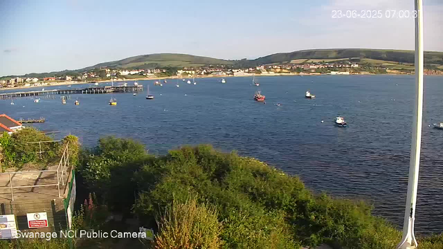 A scenic view of a body of water with sailboats and small boats floating on it, bordered by a shoreline with buildings, hills covered in greenery, and a pier extending into the water. The sky is mostly clear with some light clouds. The image is timestamped June 23, 2025, at 7:00 AM.