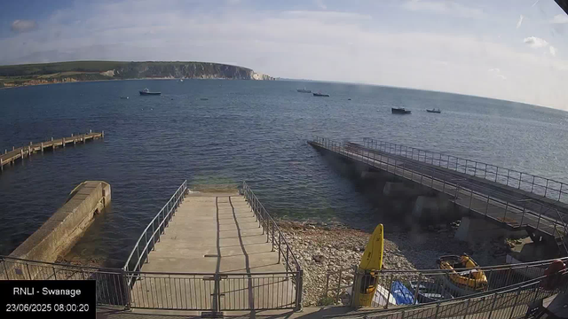A view of a harbor with a concrete and metal railing walkway leading into the water. Several boats are anchored in the sea, with cliffs visible in the background under a partly cloudy sky. There are two sloped structures extending into the water, one of which appears to be a slipway. A yellow caution sign and some small equipment are visible near the bottom right, along with a person standing close to the railing. The scene is captured in the morning, as indicated by the timestamp.