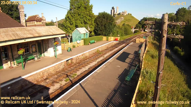 A small railway station with a platform, green benches, and a small green shed. There is a building with a sloped roof and hanging flower baskets on the left. Tracks run through the middle of the image, with a shadow cast on the platform. In the background, there are lush green trees and a hilltop with ancient ruins or castle remains. The sky is clear and sunny. There are power lines overhead and fence panels along the platform.