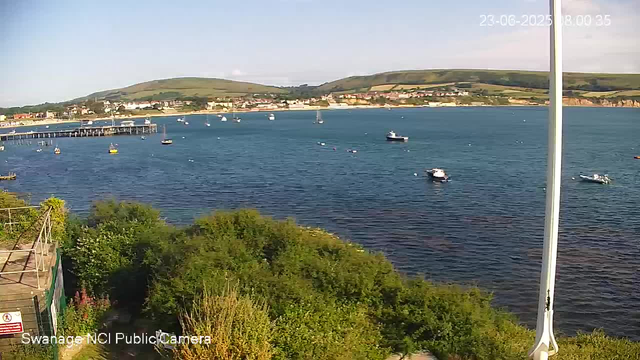 A scenic view of a harbor with several small boats floating on calm blue water, with a coastal town and rolling green hills in the background. The sky is clear with a few clouds, and the sunlight casts a warm glow over the scene. A light pole is visible on the right side of the image, and green foliage is in the foreground. The timestamp in the upper right corner indicates the date and time as June 23, 2023, at 8:00:35 AM.