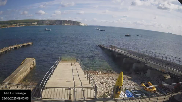 A view of a harbor with a concrete pier extending into the water. Several small boats are anchored in the sea, and a larger ship is visible in the distance. The sky is partly cloudy with patches of blue, and white cliffs or hills are visible on the horizon. In the foreground, there are safety barriers, a yellow caution sign, and some equipment near the shoreline. The timestamp indicates the date and time as 23/06/2025 at 09:00:06.
