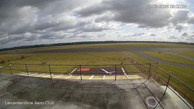 A view from a webcam showing an airport runway and surrounding grassy areas under a mostly cloudy sky. There is a helipad marked with a red square and a white helicopter symbol in the foreground, along with a small platform and guardrails. The scene is labeled "Leicestershire Aero Club" and the timestamp indicates it was taken on June 23, 2025, at 9:00 AM.