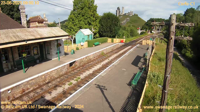 A small railway station platform with a stone building on the left, decorated with hanging flower baskets. There are green benches, a green sign, and a black lamppost casting a shadow on the platform. Beyond the platform, there are multiple railway tracks, a green shed, and yellow safety barriers. In the background, a hillside with lush green trees and a historic castle ruin on top of the hill. The sky is partly cloudy, and the scene appears to be taken in the morning.