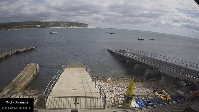 The image shows a view of a harbor with several boats floating on the water under a cloudy sky. In the foreground, there is a concrete pier extending into the water, with railings on each side, and a rocky shoreline below. To the right, a ramp leads into the water, supported by large concrete pillars. On the shoreline near the bottom right corner, there are yellow safety buoys, an inflatable boat, and some equipment. In the background, a green landmass with white cliffs is visible beyond the water.