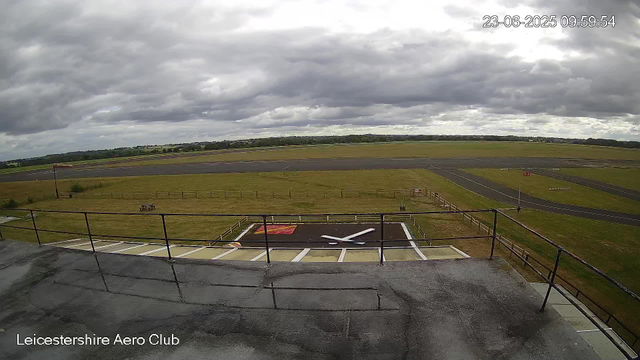 A view from a webcam overlooking a small helicopter landing pad at Leicesterhire Aero Club. The pad has a marked helicopter landing zone with a red and white logo in the middle. Beyond the pad, there is a grassy area surrounded by a fence, with an aircraft windsock on a pole. In the background, there are runways and open fields under a cloudy sky, with some distant trees and landscape. The timestamp indicates the date and time as June 23, 2025, at 09:59:54.