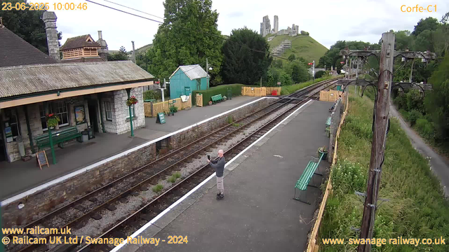 A bald man wearing a gray hoodie and shorts is standing on a railway platform, taking a photo with his phone. The platform has a stone building with hanging flower baskets and a green bench. There are some potted plants near a small teal-colored shed, and a yellow fence encloses part of the area. In the background, a green hillside with a castle ruin on top is visible, along with trees and some power lines. The weather appears overcast.
