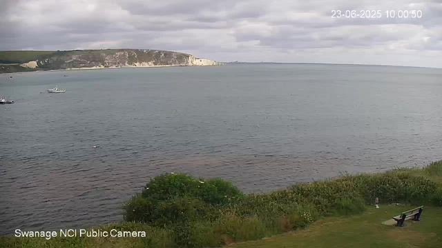 A view of a large body of water, with a shoreline of white cliffs in the distance under cloudy skies. There are a few small boats floating on the water. In the foreground, there is a grassy area with bushes and two benches, one of which has a person sitting on it. The image includes a timestamp in the top right corner and the bottom left corner has the text "Swanage NCI Public Camera."
