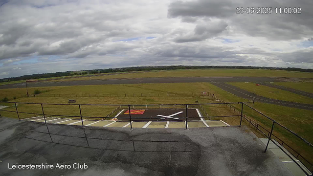 A view from a webcam at Leicesterhire Aero Club showing a cloudy sky over an airfield with runways and grassy areas. In the foreground, there is a helipad marked with a white "H" on a red background, surrounded by a black asphalt surface with painted white lines. The image has a timestamp in the upper right corner showing "23-06-2025 11:00:02".