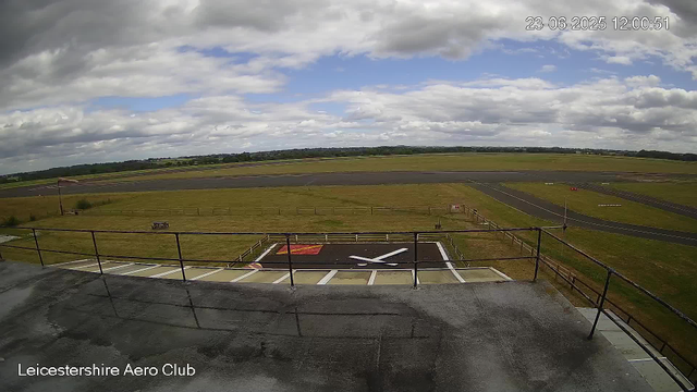 A view from a webcam showing an airfield with a helipad in the foreground. The helipad has a large white "H" and red markings, and is surrounded by safety lines and fencing. Beyond the helipad, there are grassy areas and taxiways. In the background, there are open fields and a cloudy sky. The timestamp on the image reads "23-06-2025 12:00:51," and it is labeled "Leicestershire Aero Club" at the bottom.