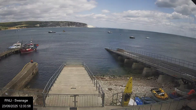 A view of a harbor with a concrete pier and boats docked. Several boats are sailing in the water, with some in the distance. The sky is partly cloudy, and there are green hills with white cliffs in the background. In the foreground, there are yellow safety equipment and a small inflatable boat inside a fenced area. The timestamp reads "23/06/2025 12:00:58," and the location is indicated as "RNLI - Swannage."