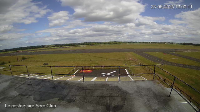 An outdoor scene at Leicestershire Aero Club showing a runway under a partly cloudy sky. In the foreground, there is a platform with a metal railing, a helicopter landing pad marked with a red square and a white helicopter icon, and a small white sign. The background features a grassy area around the runway, with fields extending into the distance and a line of trees on the horizon. The timestamp in the upper right corner indicates the date and time as June 23, 2025, at 13:00:11.