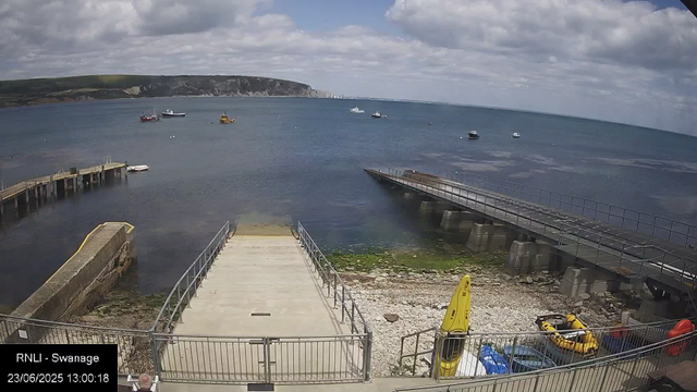 A view of the waterfront at Swanage, showing a concrete pier leading into the water, with a wheelchair-accessible ramp on the right. Several small boats are docked or anchored in the water, and a few are near the shore. In the foreground, there are yellow rescue equipment and a small orange lifeboat inside a fenced area. The background features a hilly coastline with a partly cloudy sky overhead. The timestamp indicates the date and time as June 23, 2025, at 1:00 PM.