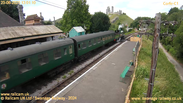 A vintage green steam train is stopped at a rural railway station platform. The steam engine is emitting a small puff of white steam. The platform has a green bench and a potted plant. Overhead electric lines are visible on a wooden post to the right. In the background, there are trees, a few small buildings, and a hill with the ruins of an old castle at the top. The sky is partly cloudy.