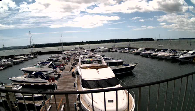 A marina filled with various boats and yachts docked along wooden piers, with calm water and a cloudy sky overhead. A curved railing is visible in the foreground.
