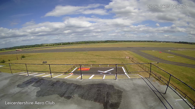 A view from a webcam at Leicesterhire Aero Club showing the tarmac runway and surrounding airfield under a partly cloudy sky. In the foreground, there is a patio area with a black surface, white parking lines, and a large painted white check mark on the ground. A small flagpole with a flag is on the left side. The airfield extends into green grassy areas with a walking fence, and distant trees and fields can be seen on the horizon. The timestamp in the upper right corner reads 23-06-2025 14:00:20.