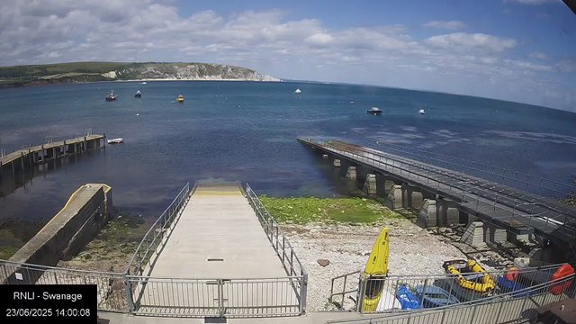 A view of a coastal area with a wooden pier extending into the water on the left and a concrete ramp on the right, leading into the sea. Several boats are floating in the water, and a large, rocky cliff extends along the horizon in the background. The sky is partly cloudy with patches of blue, and the shoreline has some green patches and rocks. In the foreground, there are safety yellow boats and equipment, as well as parked cars and a yellow caution sign. The timestamp shows "23/06/2025 14:00:08," and the location is labeled "RNLI - Swanage."