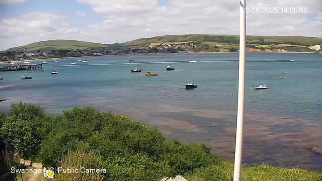 A scenic view of a bay with several anchored boats on calm waters. In the background, there are green hills and a small town along the shoreline. The sky is partly cloudy with patches of blue. There is a white pole on the right side of the image, and lush green foliage in the foreground. The timestamp indicates the date and time as June 3, 2025, at 14:00:46.
