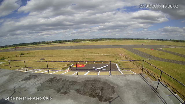 A wide view of an empty airport runway and tarmac area under a partly cloudy sky. In the foreground, there is a parking area with white painted markings and a red and white section. A fence surrounds the parking area, and a picnic table is visible on the grass beyond the fence. The landscape stretches into the distance with fields and a line of trees at the horizon. The caption in the bottom left reads "Leicestershire Aero Club." The top right corner shows the date and time: "23-06-2025 15:00:02."