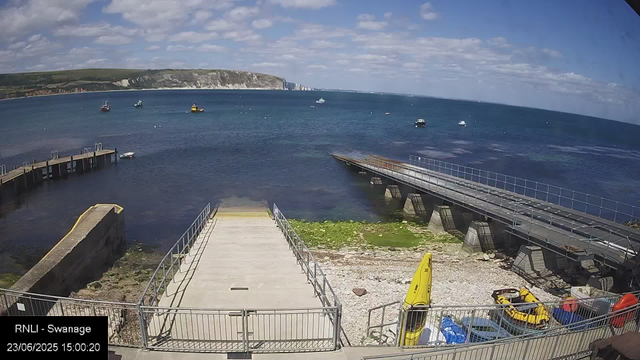 A view of the harbor at Swanage, showing a concrete entrance leading into the water with groups of yellow and black rowing boats and some cars parked nearby. Several boats are floating on the calm sea, and there is a towering white cliff with patches of greenery on the horizon under a partly cloudy sky.