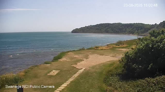 A scenic view of a coastal area with a sandy beach, a grassy path with stepping stones leading toward the water, and a lush, green hill in the background under a clear sky. The sea is calm with gentle waves, and the image is captured during daytime. The timestamp in the upper right corner indicates the date and time as June 23, 2025, at 3:00 PM.
