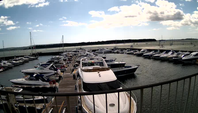 A marina with multiple boats docked along wooden piers. The boats vary in size and style, some with covers and others open. The water is calm, and the sky above is partly cloudy with patches of sunlight. In the distance, there is a dark landmass or shoreline. The image is taken from a vantage point behind a curved metal railing.