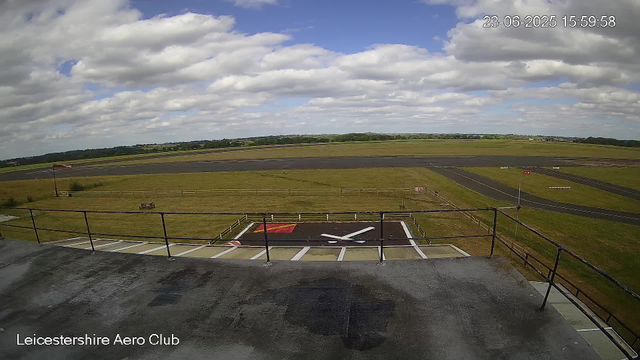 The image shows a view of an airport runway and surrounding grassy fields under a partly cloudy sky. In the foreground, there is a helipad with a large white "H" and a red square marking. A metal railing surrounds the edge of the structure from which the photo was taken. To the left, there is a small flagpole with a flag fluttering in the wind, and a bench nearby. The background features a flat landscape with open fields and distant trees, and the scene appears to be captured from a high vantage point at Leicestershire Aero Club. The date and time are displayed in the upper right corner as June 23, 2025, at 15:59:58.