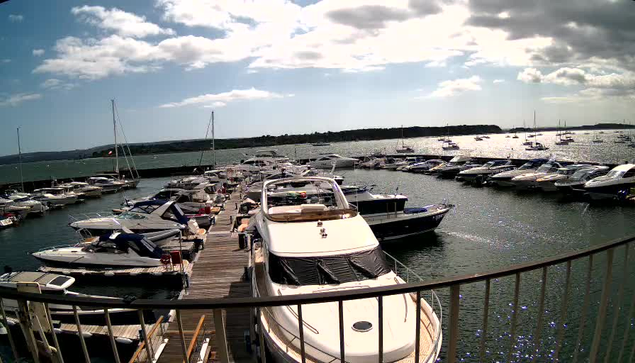 A marina filled with numerous boats and yachts docked along wooden piers. The water is calm, and the sky is partly cloudy with patches of blue and sunlight shining through. A curved railing is visible in the foreground, indicating the photo was taken from a viewpoint above the dock.