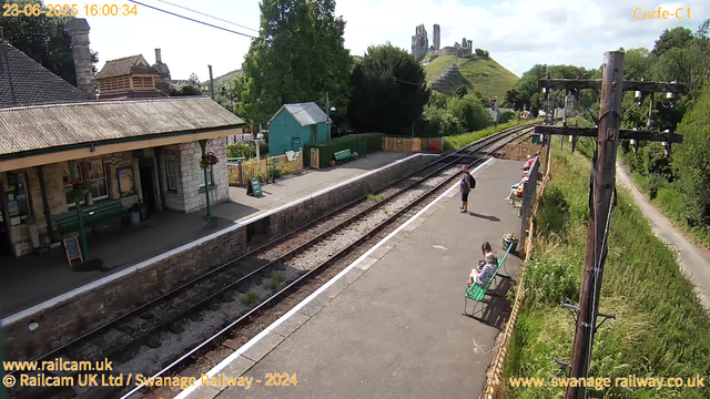 A small train station platform with a stone building on the left and a green bench in front of it. Several people are sitting on benches along the right side of the platform, while a man stands near the tracks. Overhead, there are tall wooden electrical poles with wires. In the background, a green hill with ruins or castle remains on top is visible, along with trees and a partly cloudy sky. The tracks run through the center of the image, with a narrow path and grassy area on the right.