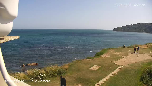 A view of the ocean with a small grassy and sandy area in the foreground. Two people are walking along the path, and a small dog is nearby. There is a rocky headland on the right side in the distance, and a sailboat is visible far out on the water. The sky is clear and blue. The timestamp in the top right corner reads "23-06-2025 16:00:58."