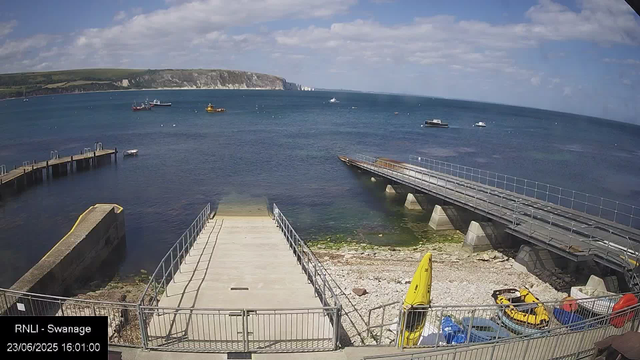 A clear day at the harbor shows a small beach area with a concrete pier extending into the water. Several boats are anchored in the bay, including small boats and larger vessels. There is a yellow kayak and some other small watercraft on the beach, along with safety equipment like a yellow lifeguard’s float. A railing surrounds the beach area, which has some rocky and gravel patches, and the sea stretches out under a partly cloudy sky with white fluffy clouds. In the background, there are white cliffs and green land at a distance.