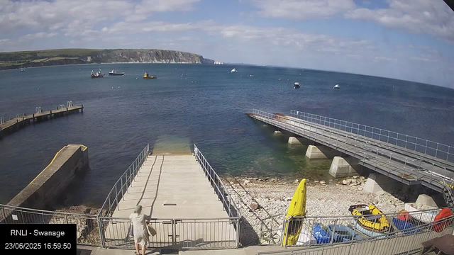 A view of a seaside harbor with a concrete pier extending into calm blue water. Several boats are anchored offshore, and there is a hilly green landscape in the background under a partly cloudy sky. In the foreground, a person is walking down a ramp toward the water, with some small vehicles and a yellow surfboard or paddleboard leaning against the fence. The timestamp indicates the photo was taken on June 23, 2025, at 4:59 PM.