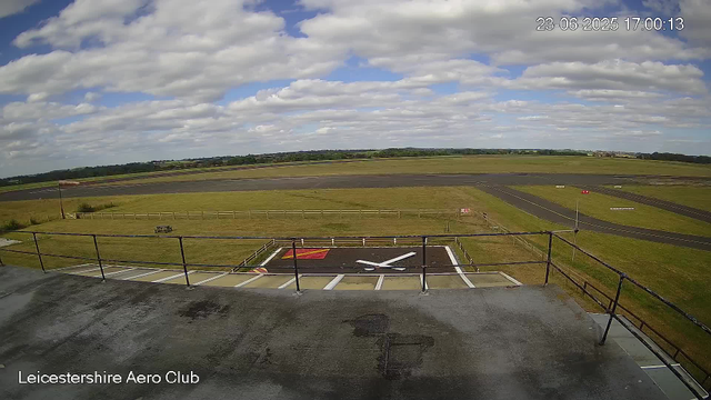 A view from a webcam at Leicestershire Aero Club showing the airfield under a partly cloudy sky. In the foreground, there is a helicopter pad with a white rotor symbol and a red and yellow marking. The field is surrounded by grassy areas and the runways extend into the distance. The image includes a timestamp showing the date as June 23, 2025, at 17:00:13.