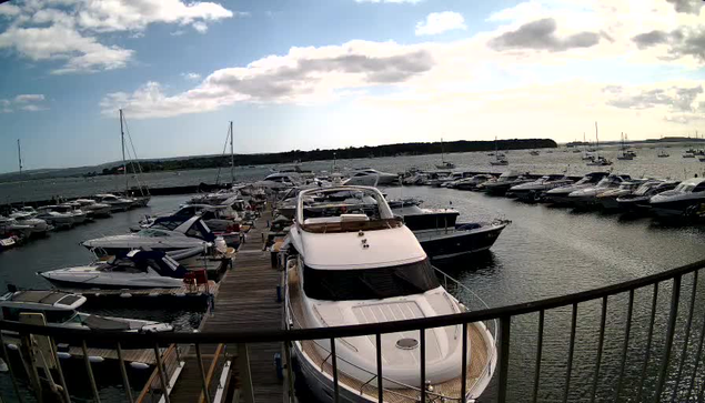 A marina filled with numerous boats and yachts docked along wooden piers under a partly cloudy sky, with some boats anchored further out in the water and a distant shoreline visible on the horizon.