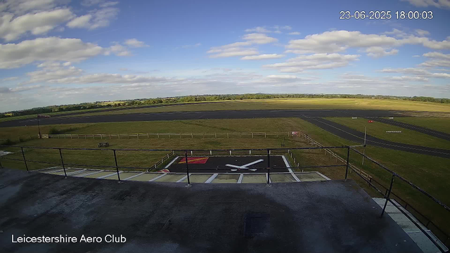 A view from a webcam at Leicestershire Aero Club shows a clear sky with scattered clouds, a runway stretching across the landscape, a helicopter landing pad with a white "H" and an aircraft symbol marked on it in the foreground, and lush green fields in the background. The timestamp reads 23-06-2025 18:00:03.
