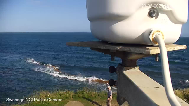 A webcam captures a view of the ocean with clear blue sky above. In the foreground, a large white weather instrument or sensor is mounted on a wooden platform. Below, near the shoreline, a person wearing a wide-brimmed hat, a blue shirt, and beige pants stands on a grassy area, looking toward the water.