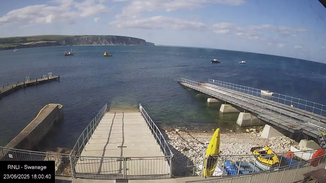 A view of a harbor with a concrete pier extending into calm blue water. Several small boats are floating in the distance, and a few are docked or near the pier. There is a yellow kayak and some equipment on the shoreline, and a fenced-off area with a ramp leading into the water. In the background, green cliffs rise against a partly cloudy sky.